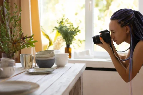 Fotógrafa usando uma câmera para fotografar louças, copos e outros objetos organizados sobre uma mesa, representando a produção de fotos de produtos para vendas no e-commerce