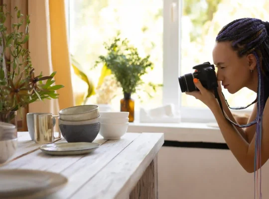 Fotógrafa usando uma câmera para fotografar louças, copos e outros objetos organizados sobre uma mesa, representando a produção de fotos de produtos para vendas no e-commerce