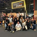 Equipe da JoomPulse reunida em frente ao estande da empresa durante um evento de e-commerce, com o logotipo da JoomPulse iluminado acima do grupo
