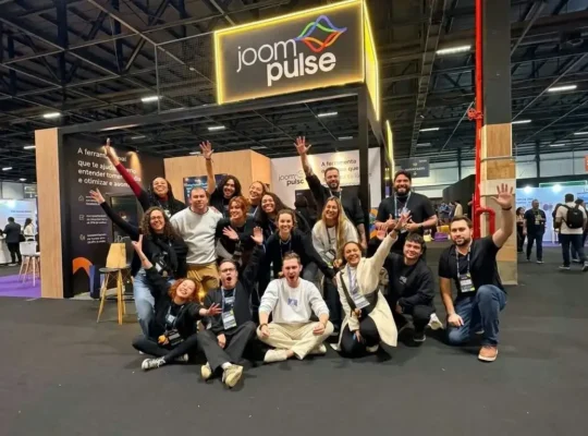 Equipe da JoomPulse reunida em frente ao estande da empresa durante um evento de e-commerce, com o logotipo da JoomPulse iluminado acima do grupo