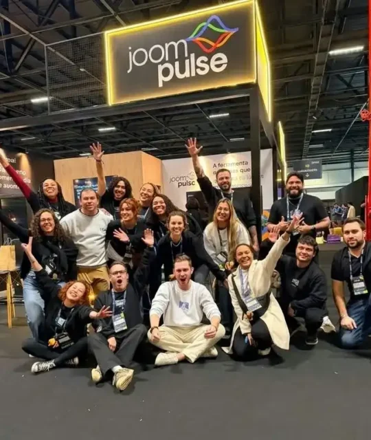 Equipe da JoomPulse reunida em frente ao estande da empresa durante um evento de e-commerce, com o logotipo da JoomPulse iluminado acima do grupo