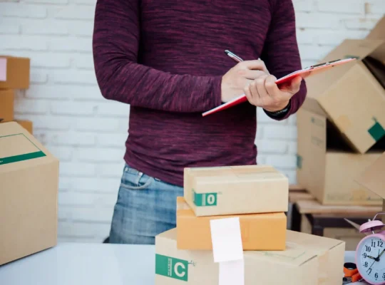 Pessoa conferindo pedidos em uma prancheta ao lado de várias caixas de produtos prontas para envio