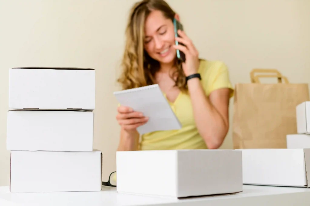 Vendedora falando ao celular enquanto confere anotações, cercada por caixas de produtos e embalagens, representando a escolha e preparação de produtos para vender na Shopee
