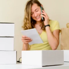 Vendedora falando ao celular enquanto confere anotações, cercada por caixas de produtos e embalagens, representando a escolha e preparação de produtos para vender na Shopee