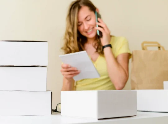 Vendedora falando ao celular enquanto confere anotações, cercada por caixas de produtos e embalagens, representando a escolha e preparação de produtos para vender na Shopee