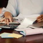 Pessoa usando calculadora ao lado de recibos, documentos e notebook
