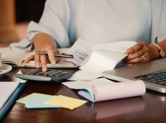 Pessoa usando calculadora ao lado de recibos, documentos e notebook