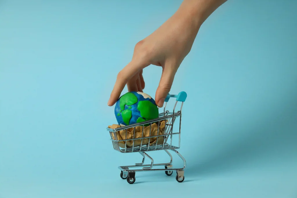 Mão colocando um pequeno globo terrestre dentro de um carrinho de compras em miniatura sobre fundo azul
