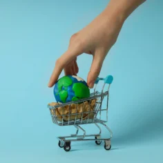 Mão colocando um pequeno globo terrestre dentro de um carrinho de compras em miniatura sobre fundo azul