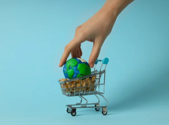 Mão colocando um pequeno globo terrestre dentro de um carrinho de compras em miniatura sobre fundo azul