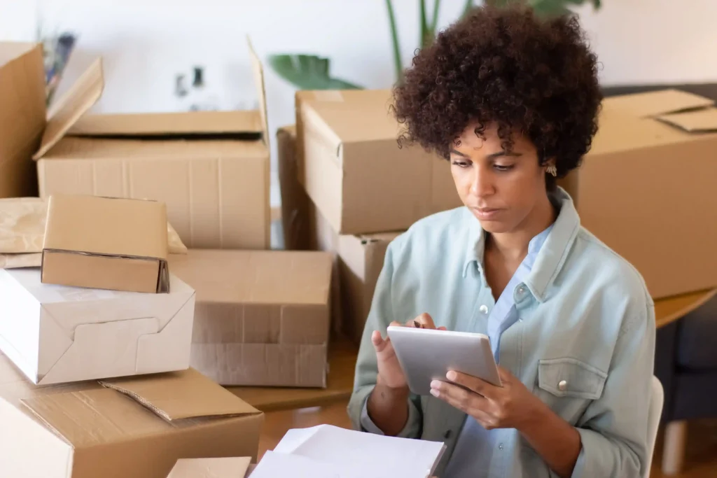 Mulher analisa pedidos em um tablet cercada por caixas de produtos para organizar o estoque antes da Black Friday 2026