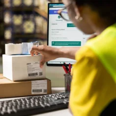 Profissional de logística organizando caixas de encomendas com etiquetas de envio, diante de um computador com informações de pedidos e entrega