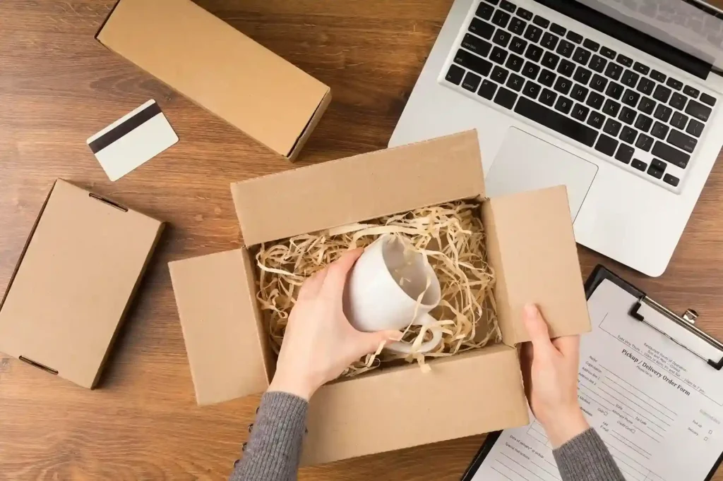 Vista superior de uma pessoa colocando uma caneca branca em uma caixa de papelão com enchimento de papel, sobre uma mesa com notebook, outras embalagens e formulário de entrega