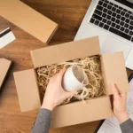 Vista superior de uma pessoa colocando uma caneca branca em uma caixa de papelão com enchimento de papel, sobre uma mesa com notebook, outras embalagens e formulário de entrega