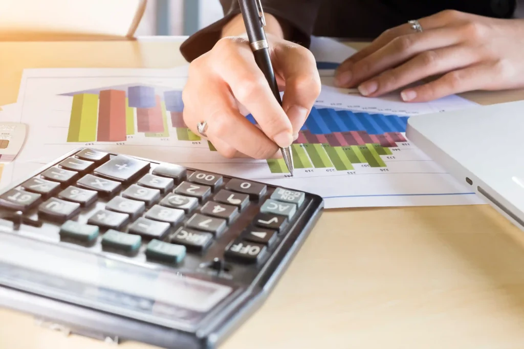 Pessoa utilizando uma caneta para analisar gráficos financeiros coloridos sobre uma mesa, com calculadora e notebook ao lado