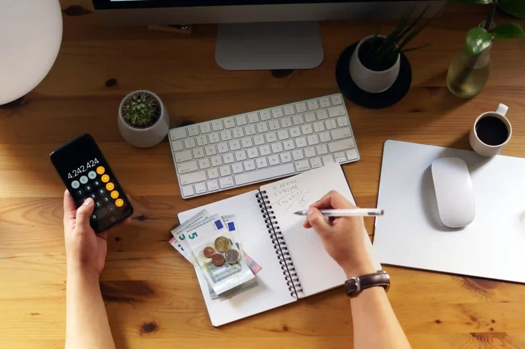 Pessoa utilizando a calculadora do celular enquanto anota valores em um caderno, com notas e moedas de euro sobre uma mesa de trabalho