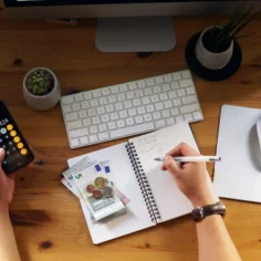 Pessoa utilizando a calculadora do celular enquanto anota valores em um caderno, com notas e moedas de euro sobre uma mesa de trabalho