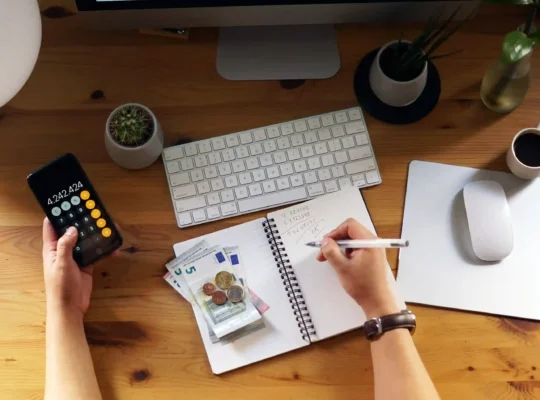 Pessoa utilizando a calculadora do celular enquanto anota valores em um caderno, com notas e moedas de euro sobre uma mesa de trabalho