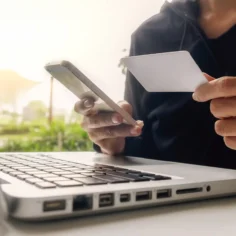 Pessoa segurando um cartão bancário e utilizando um smartphone diante de um notebook, representando os pagamentos online