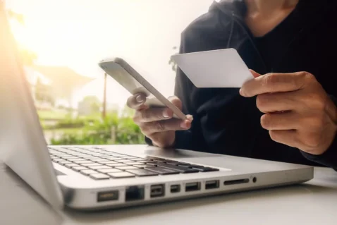 Pessoa segurando um cartão bancário e utilizando um smartphone diante de um notebook, representando os pagamentos online