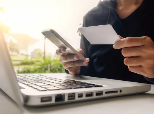 Pessoa segurando um cartão bancário e utilizando um smartphone diante de um notebook, representando os pagamentos online