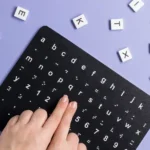 Mãos sobre uma tabela com letras e símbolos em braile, cercada por peças com letras em fundo lilás
