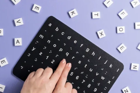 Mãos sobre uma tabela com letras e símbolos em braile, cercada por peças com letras em fundo lilás