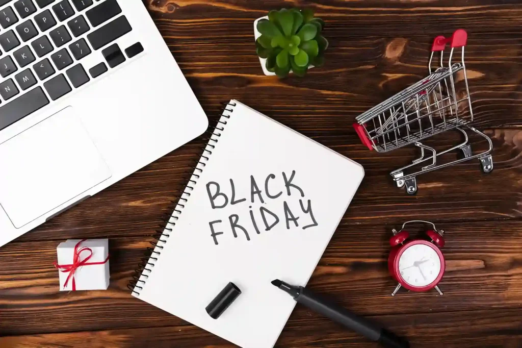 Caderno com a inscrição Black Friday sobre mesa de madeira, ao lado de notebook, carrinho de compras, presente e despertador vermelho