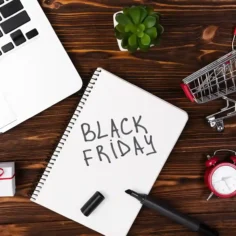 Caderno com a inscrição Black Friday sobre mesa de madeira, ao lado de notebook, carrinho de compras, presente e despertador vermelho
