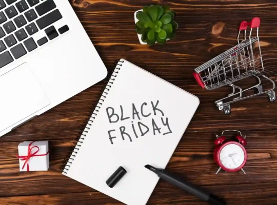 Caderno com a inscrição Black Friday sobre mesa de madeira, ao lado de notebook, carrinho de compras, presente e despertador vermelho