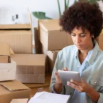 Mulher utilizando tablet para conferir informações em meio a caixas de papelão