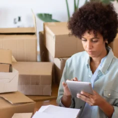 Mulher utilizando tablet para conferir informações em meio a caixas de papelão