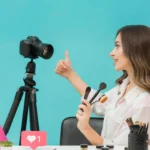Influenciadora segurando pincéis de maquiagem e fazendo sinal positivo diante de uma câmera, com ícone de curtida e fundo azul