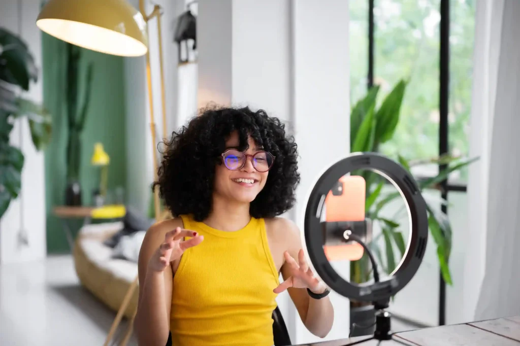 Influenciadora de óculos e blusa amarela falando diante de um celular com ring light durante a gravação de conteúdo digital