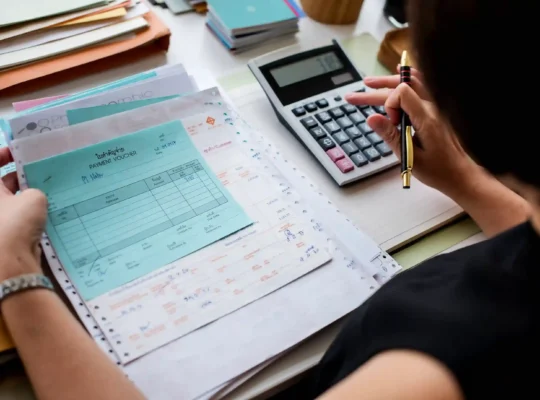 Pessoa conferindo documentos financeiros e comprovantes de pagamento enquanto utiliza uma calculadora e uma caneta sobre a mesa
