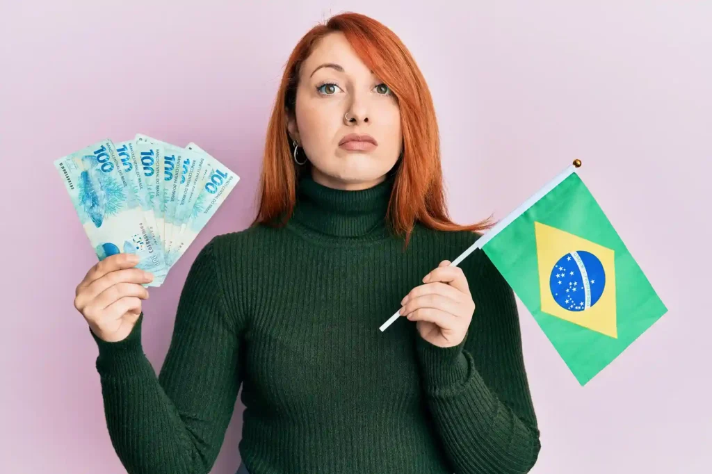 Mulher de cabelos ruivos segurando notas de 100 reais em uma mão e uma bandeira do Brasil na outra, diante de um fundo rosa-claro