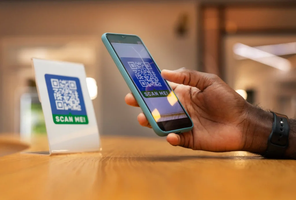 Pessoa segurando celular com código QR na tela, próximo a uma placa com código QR sobre uma mesa