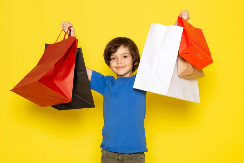 Criança com camiseta azul segurando várias sacolas de compras coloridas diante de um fundo amarelo