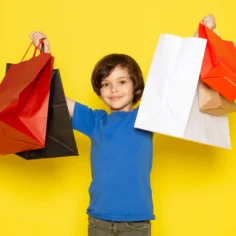 Criança com camiseta azul segurando várias sacolas de compras coloridas diante de um fundo amarelo