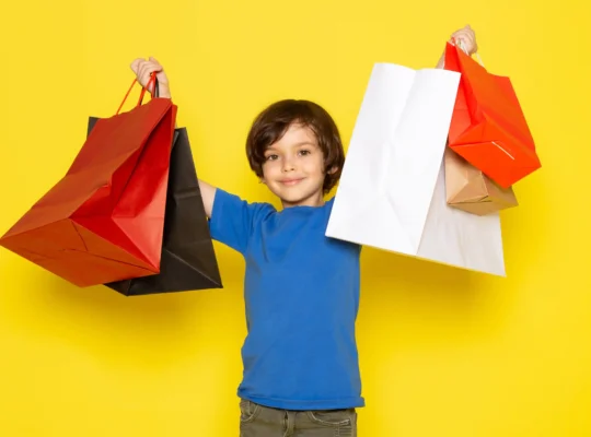Criança com camiseta azul segurando várias sacolas de compras coloridas diante de um fundo amarelo